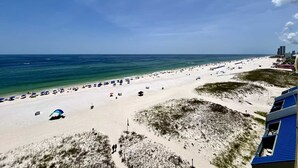 Une plage de sable blanc, avec une eau d’un bleu limpide et quelques parasols. On aperçoit des bâtiments au loin.