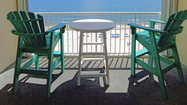 Un balcon avec deux fauteuils Adirondack verts, une petite table ronde et un ventilateur de plafond. La vue comprend une plage et l’océan.