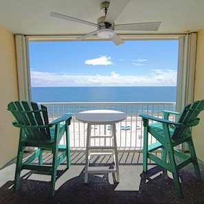 A balcony with two green Adirondack chairs, a small round table, and a ceiling fan. The view includes a beach and the ocean.