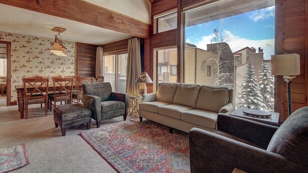 Un salon chaleureux avec un plafond en bois, un canapé, des fauteuils, une table basse et une table à manger avec des chaises. De grandes fenêtres offrent une vue sur l’extérieur.