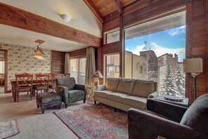Relax in this inviting living room with a high ceiling, exposed beams, and a large window offering a view of the surrounding neighborhood.