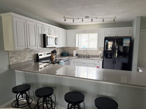 A modern kitchen with white cabinets, a stainless steel refrigerator, a large island with bar stools, and a window with blinds. - Recently-Remodeled Destin Townhouse. Extended Stay Discount Nov-Feb (Miramar Beach)