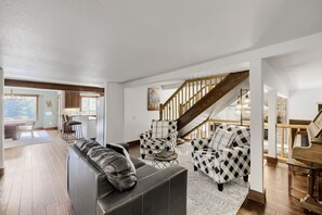 A spacious living room with a leather sofa, a checkered armchair, and a piano. A staircase with wooden railings leads to an upper level.