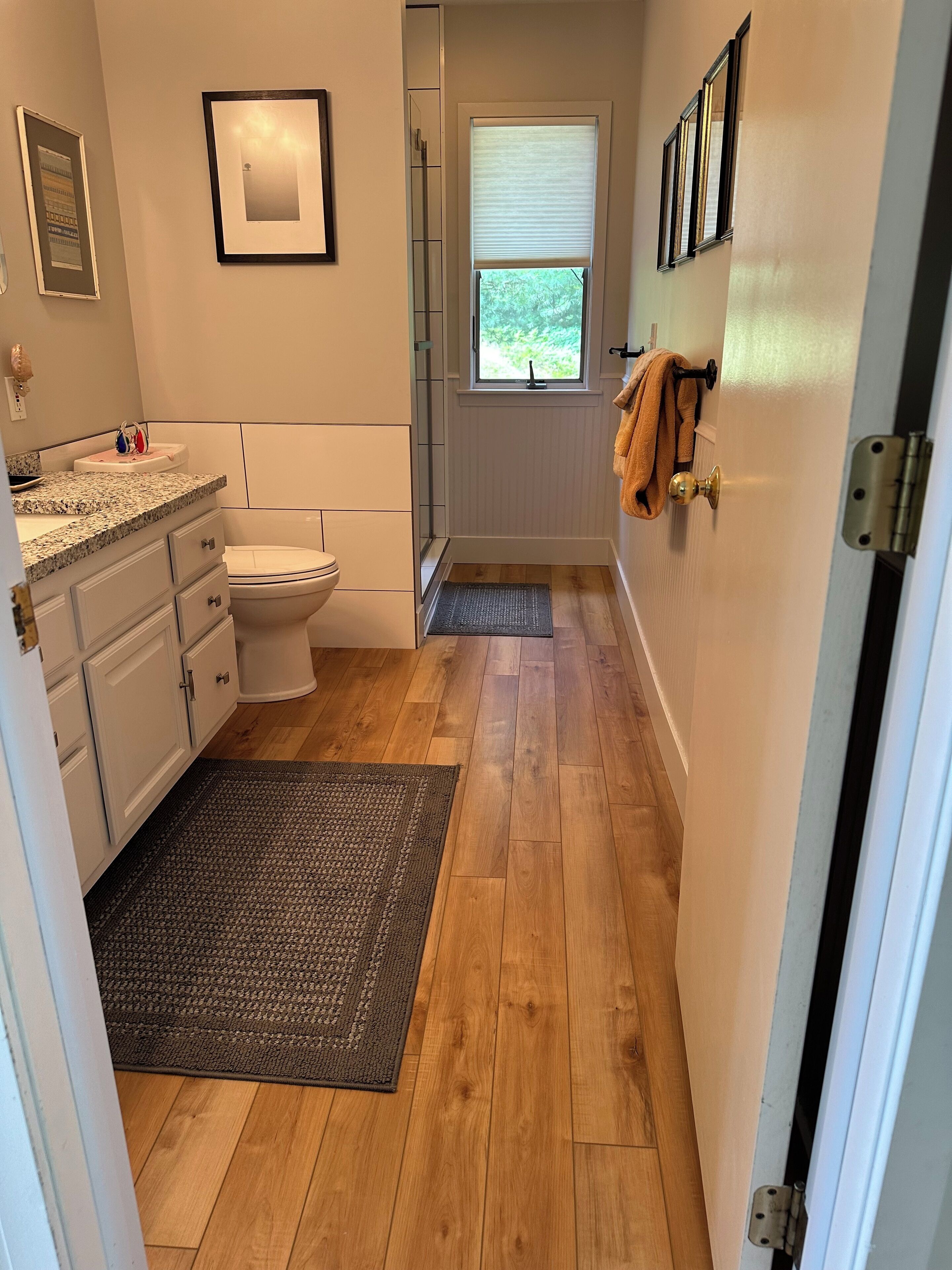 Een smalle badkamer met toilet, een wastafel met een granieten aanrechtblad, een klein raam met uitzicht op groen en een deur met een handdoek aan de hendel.