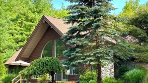 Une maison avec un toit en pente, entourée d ’un jardin bien entretenu où poussent différentes espèces de plantes et un grand arbre à feuilles persistantes.