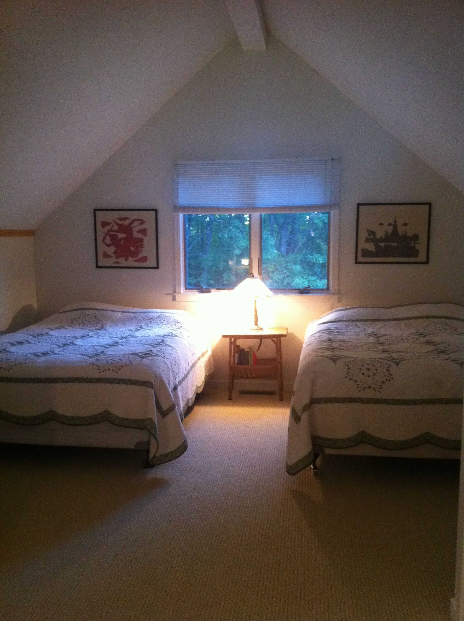 A bedroom with two beds, a nightstand, a window with blinds, and two framed pictures on the walls.