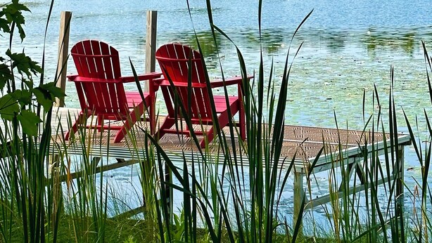 Une vue paisible sur un lac, avec deux chaises Adirondack rouges sur une jetée en bois, le tout entouré d’une végétation luxuriante et d’un lac calme.