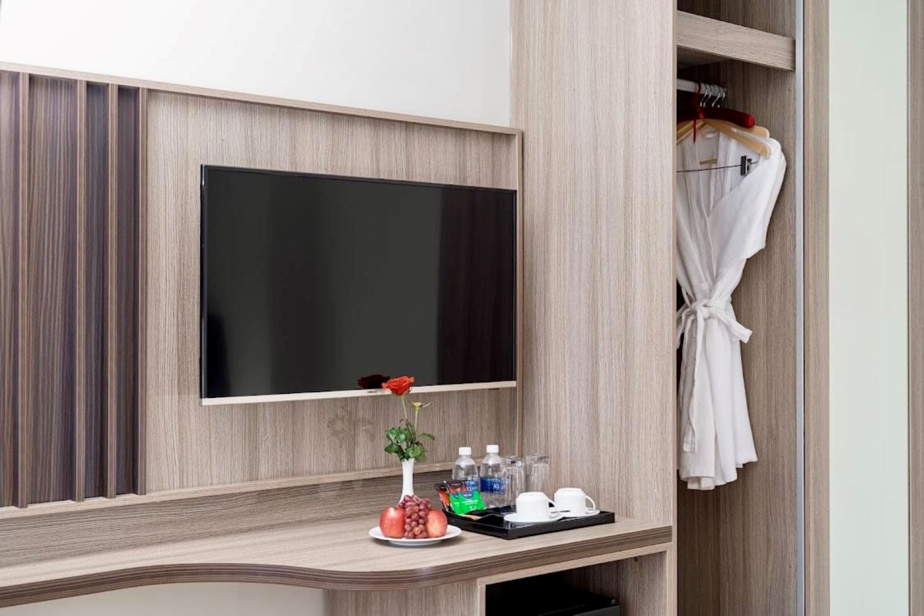 Habitación de hotel con televisión de pantalla plana en la pared, tocador con espejo, bata colgada en un perchero y bandeja con un florero, un plato de fruta y una taza.