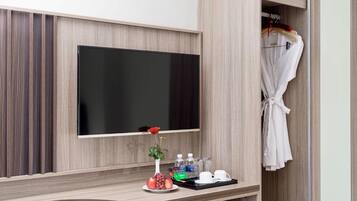 Habitación de hotel con televisión de pantalla plana en la pared, tocador con espejo, bata colgada en un perchero y bandeja con un florero, un plato de fruta y una taza.