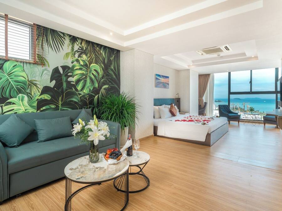 Amplia sala de estar con sofá, una mesita de centro y un gran mural tropical. En el fondo, se ve una cama con vistas al mar.