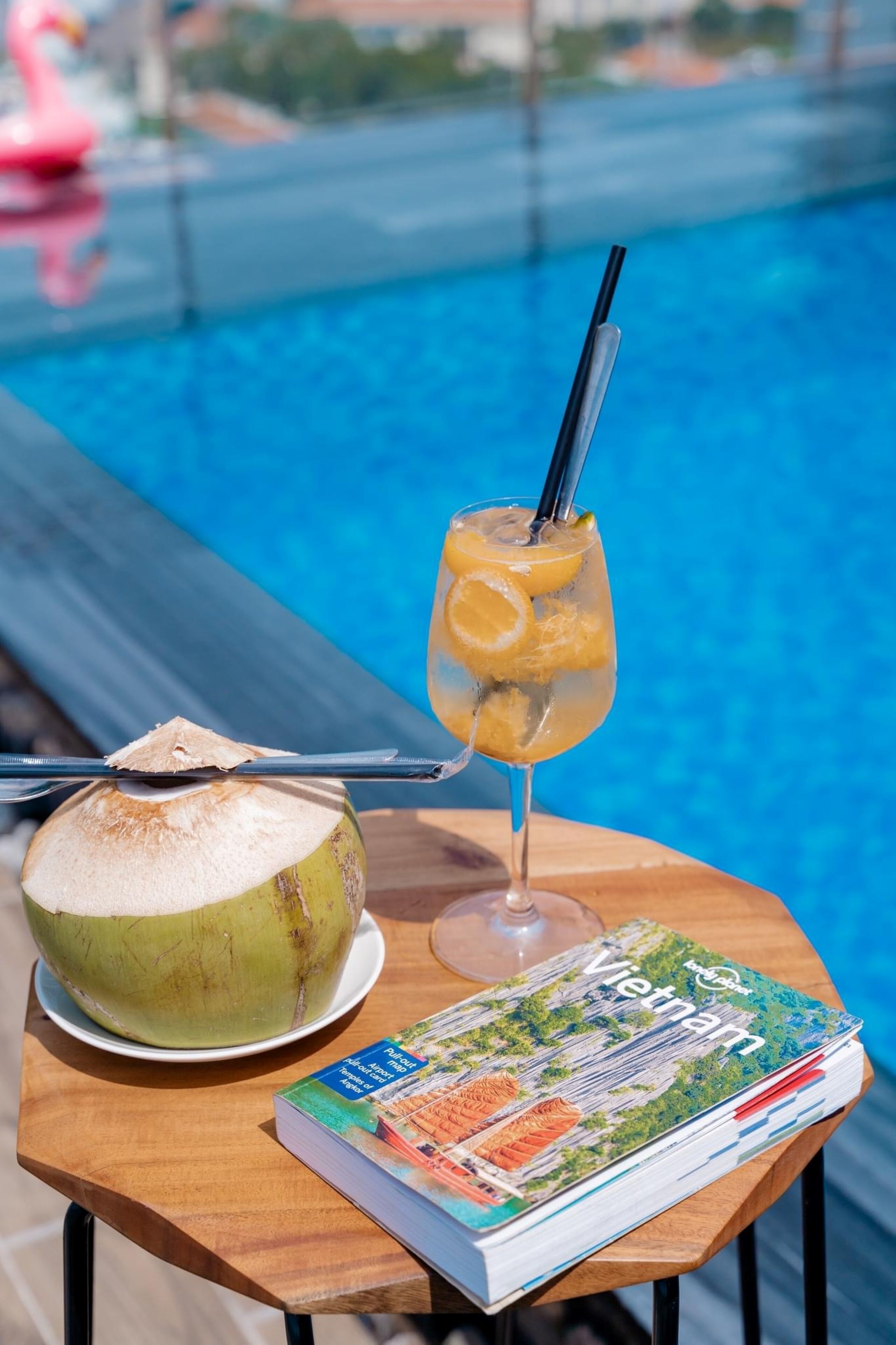 A glass of tropical drink with a straw and a coconut on a wooden table by a pool.