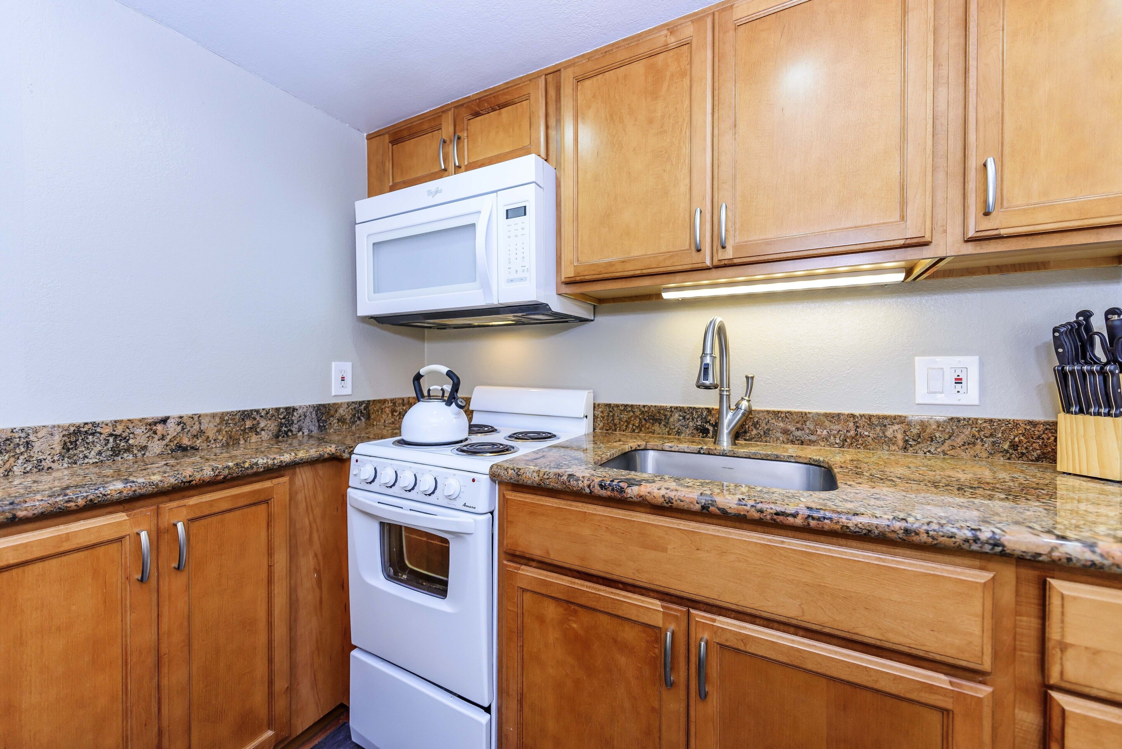 A kitchen with wooden cabinets, a white stove, a microwave, a granite countertop, and a stainless steel sink.