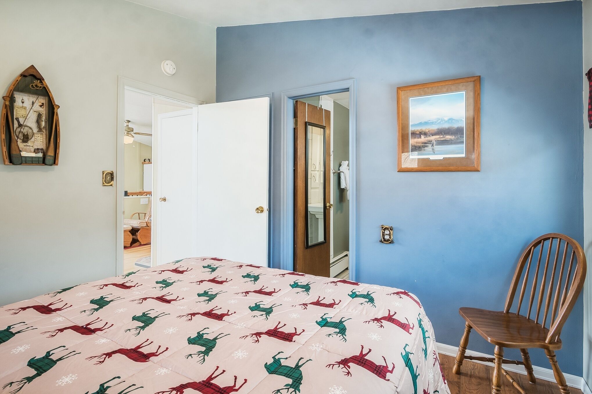 Un dormitorio con una cama, una silla de madera, una puerta que da a otra habitación y un cuadro enmarcado en la pared.