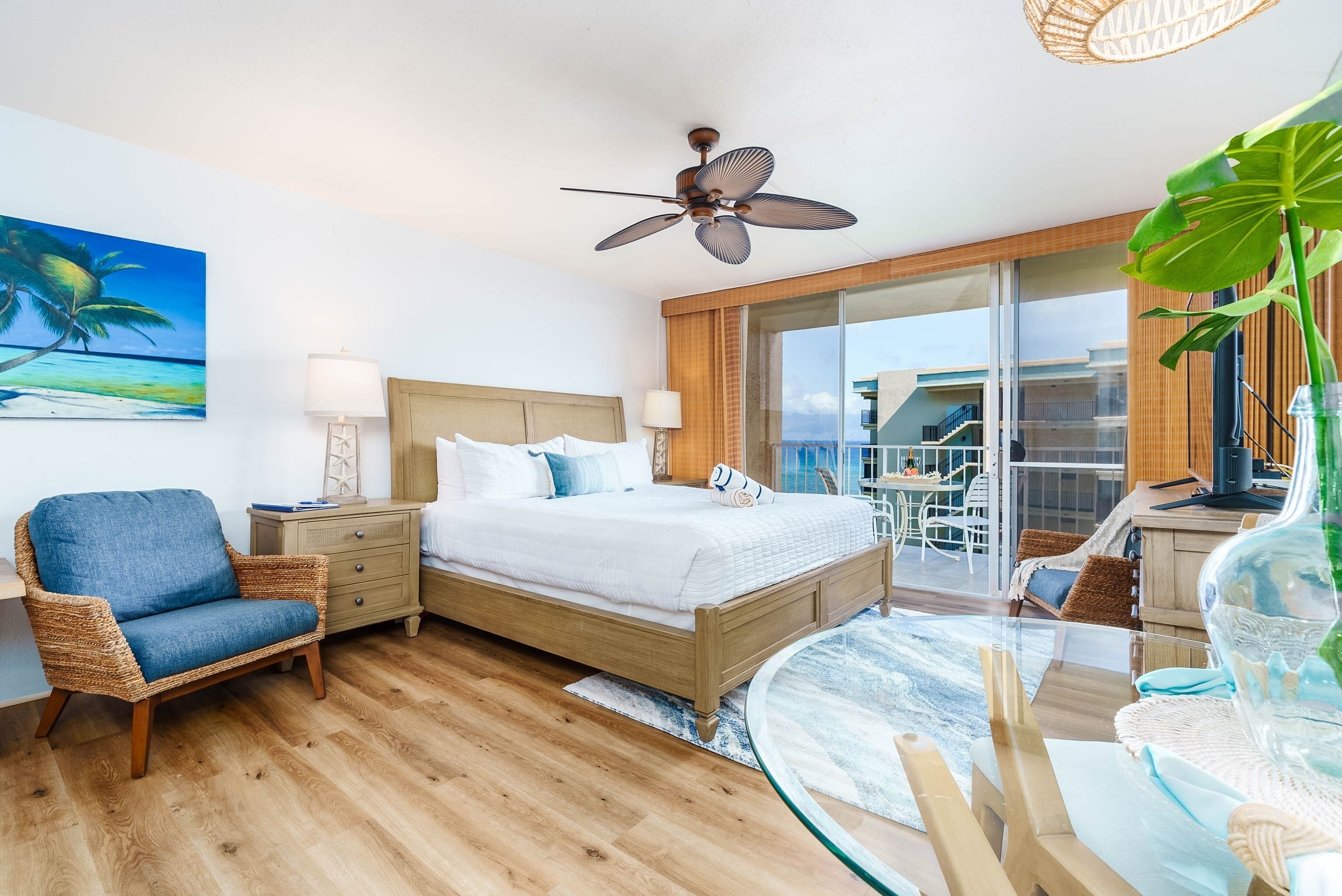 Quarto de hotel com cama, cadeira de vime, mesa de vidro, ventilador de teto e uma janela grande com vista para o exterior.
