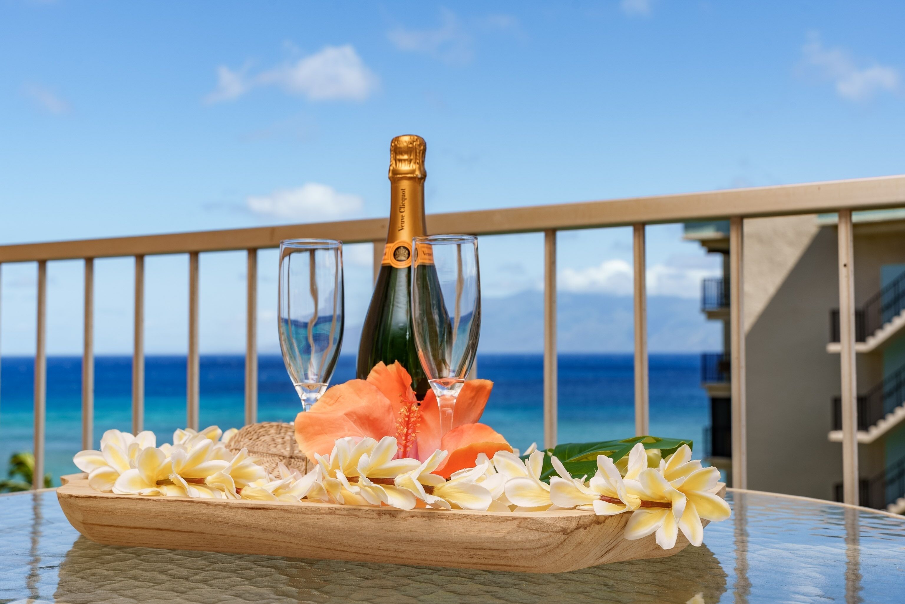 A table with a bottle of champagne, two glasses, and a wooden tray with flowers, set against a balcony with a view of the ocean.