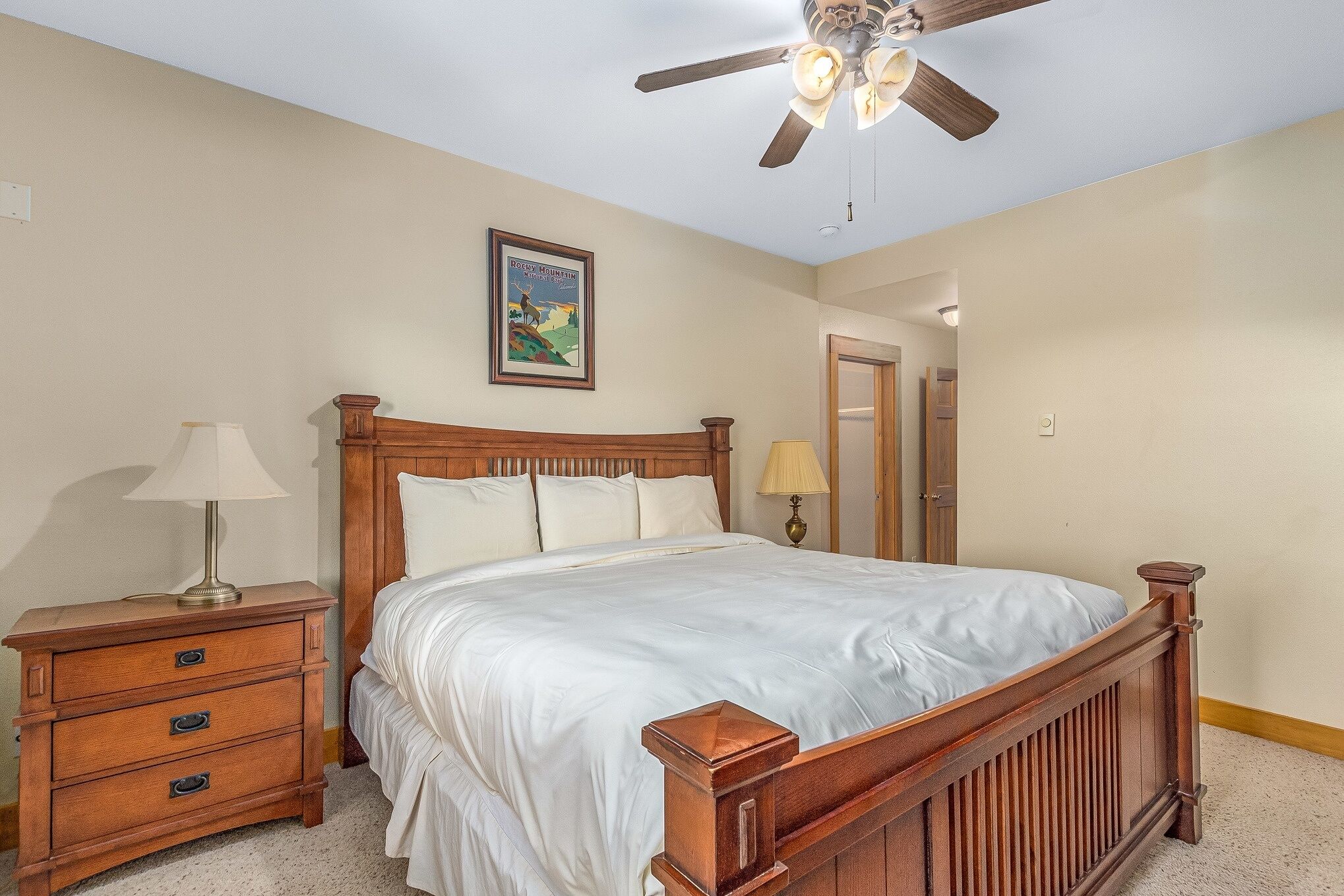 A bedroom with a wooden bed, a nightstand with a lamp, a ceiling fan, and a framed picture on the wall.