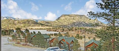 A view of a parking lot with several vehicles, surrounded by buildings with wooden exteriors and green roofs, set against a backdrop of mountains and a lake.