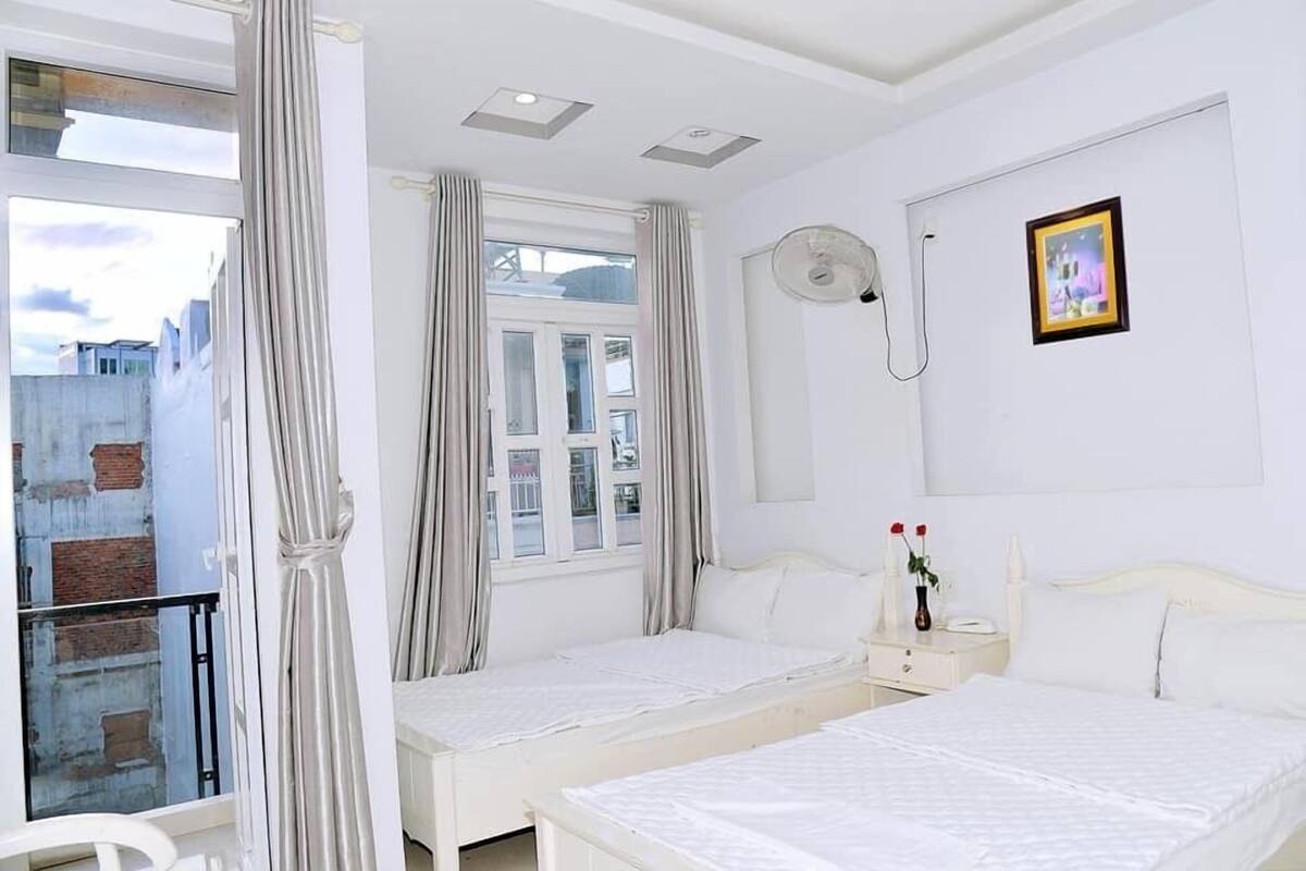 A hotel room with two beds, a balcony, a window with curtains, a bedside table with a lamp, and a picture on the wall.