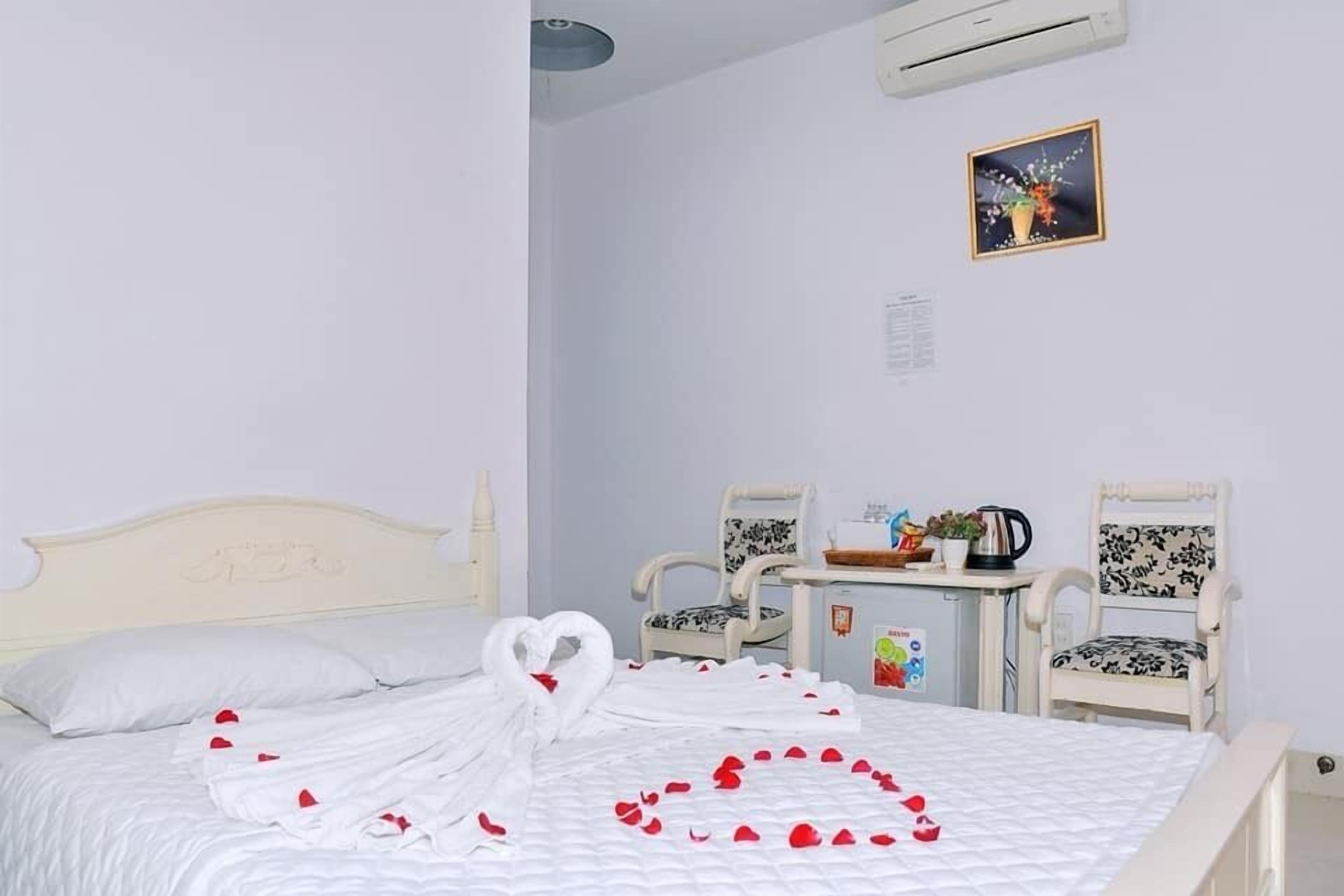 A hotel room with a bed, two chairs, a small table with a kettle and a plant, and a picture on the wall.