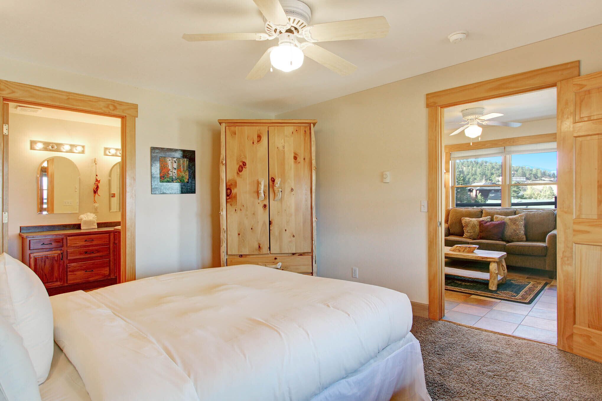 A bedroom with a large bed, wooden wardrobe, a mirror with lights, a ceiling fan, and a view of a balcony with a sofa.