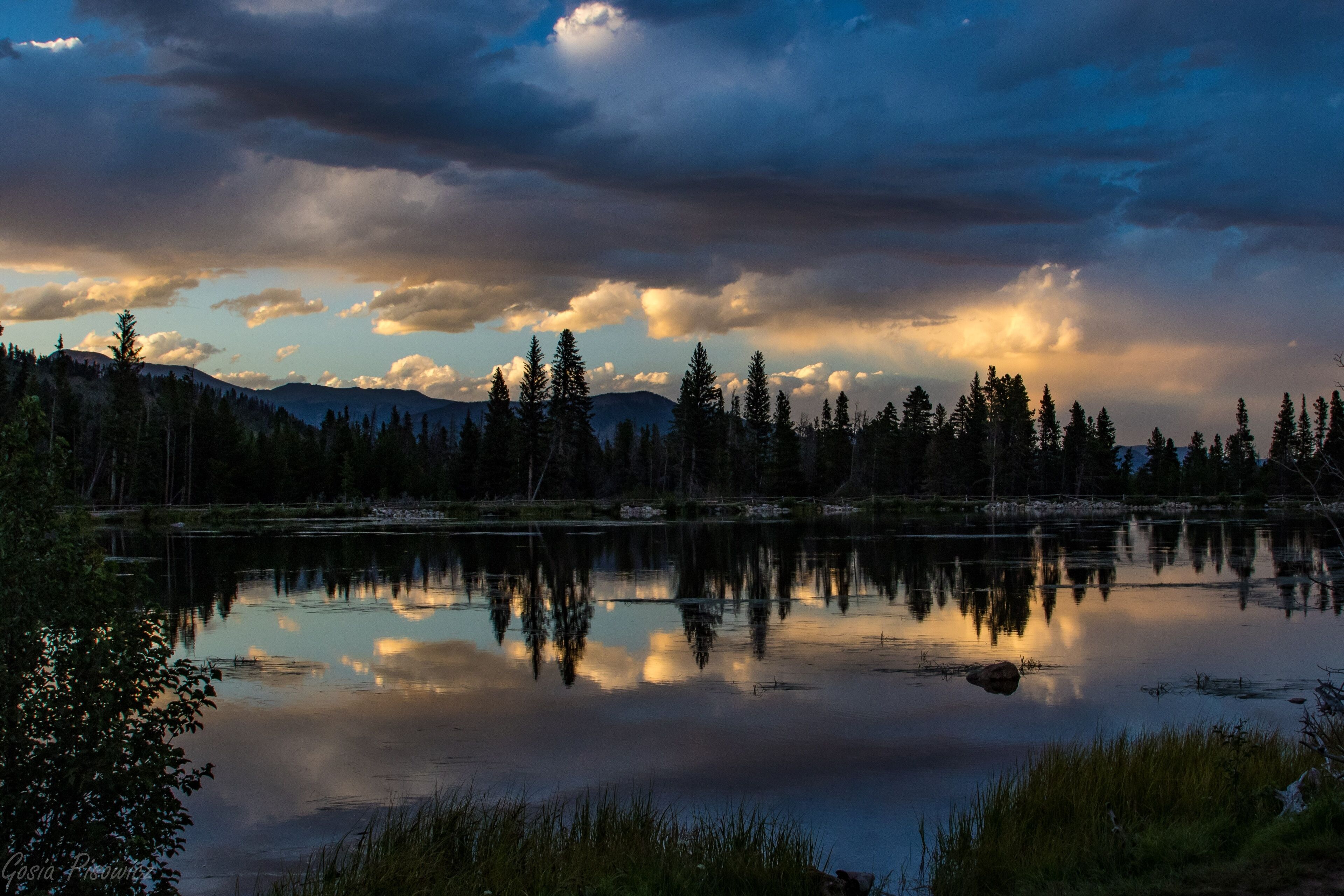 A serene lake surrounded by dense forest and mountains, with reflections of the sky and clouds on the water's surface.