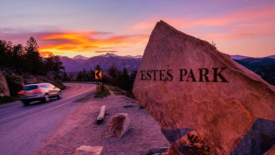 Señal de carretera para Estes Park con montañas al fondo y un atardecer.