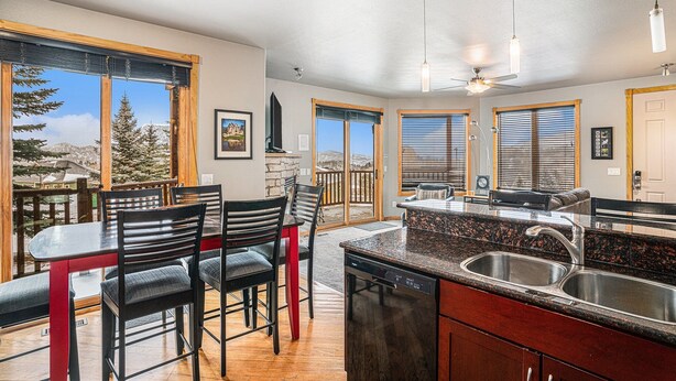 A modern kitchen with a granite countertop, stainless steel sink, and a dishwasher. There are two black chairs with red legs, a dining table, and a ceiling fan. Large windows offer a view of trees and mountains. - Nakai Peak 41b 2 Bedroom Condo by RedAwning (Estes Park)
