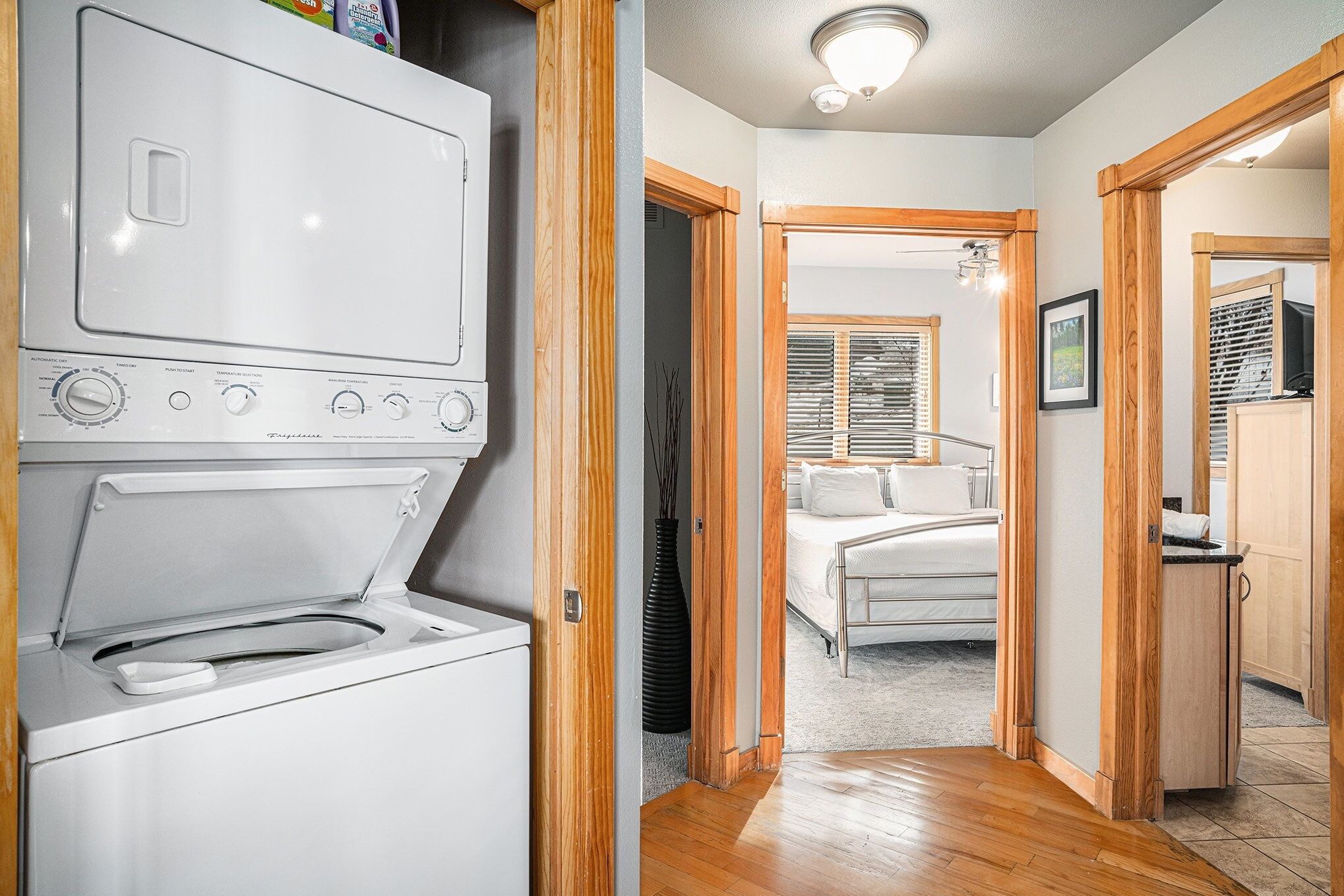 A laundry area with a front-loading washer and dryer, wooden doors leading to a bedroom with a bed and a window.