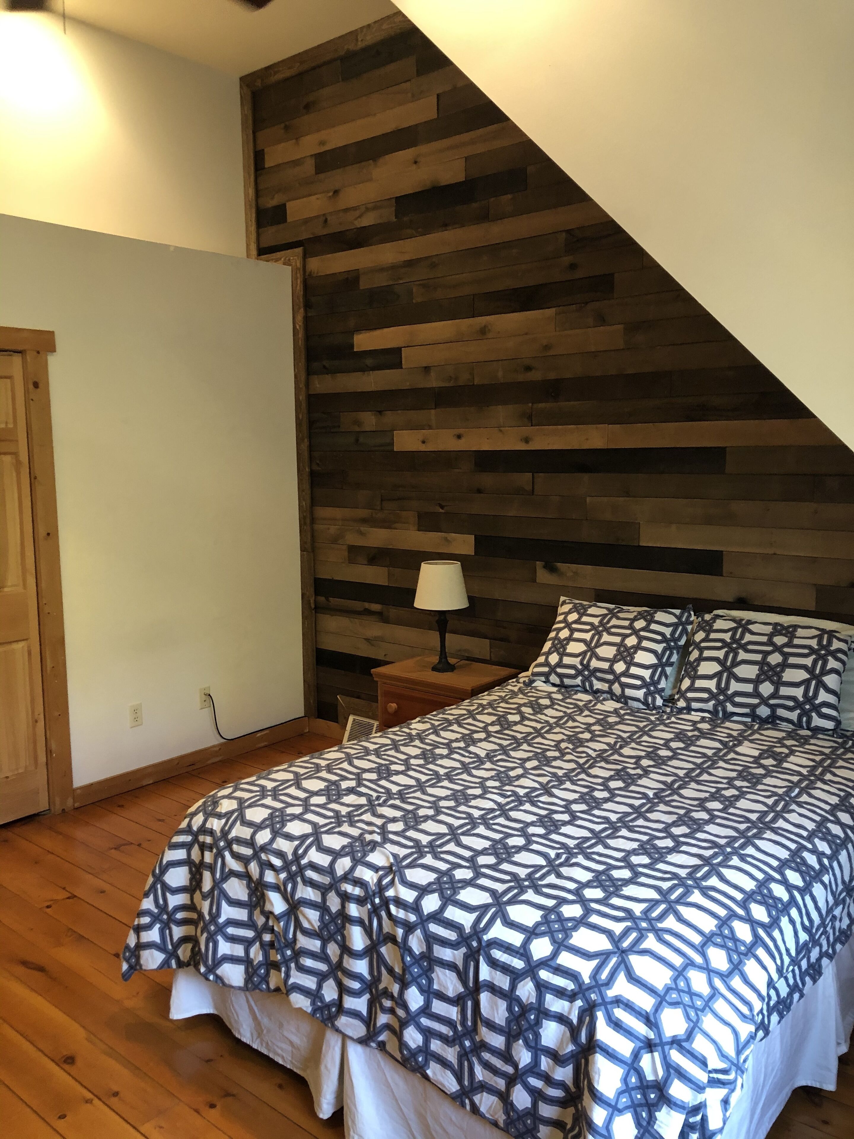 Un dormitorio con pared de listones de madera, una cama con ropa de cama estampada, una mesita de noche con lámpara y una puerta de madera.