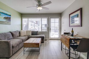 Ein modernes Wohnzimmer mit einem Ecksofa, einem Holztisch, einem Esstisch mit Stühlen, einem Deckenventilator und großen Fenstern mit Jalousien.