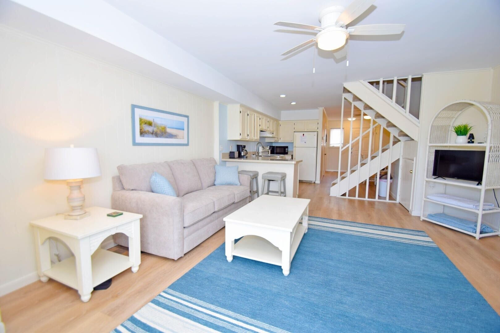 A modern living room with a sofa, coffee table, and a TV stand. There is a kitchen area with a microwave and a refrigerator. A staircase leads to an upper level.