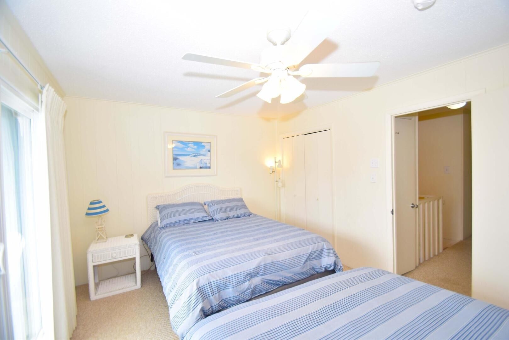 Een slaapkamer met een bed, nachtkastje, plafondventilator, een wandlamp en een deur die naar een andere kamer leidt.