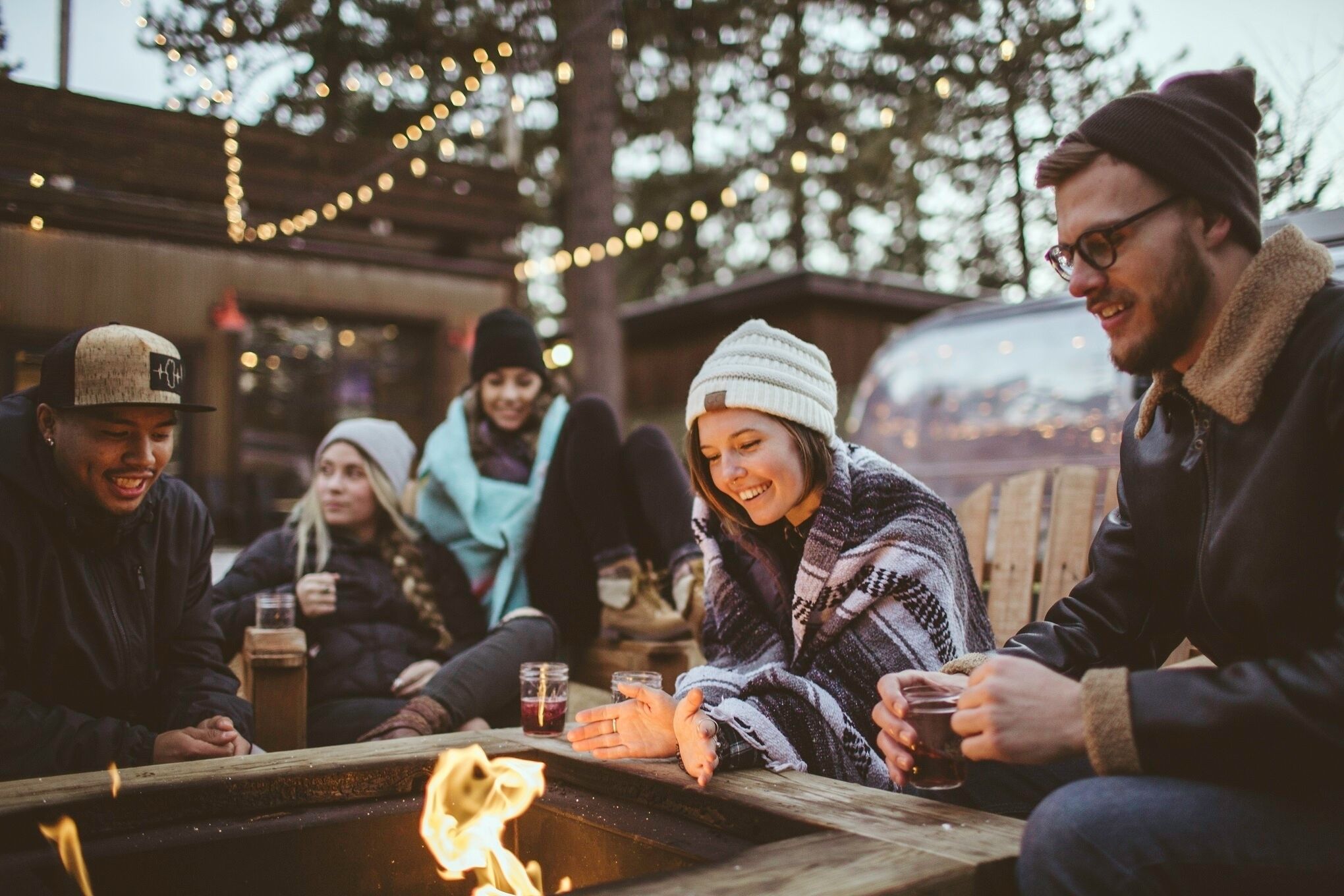 A group of people gathered around a campfire in an outdoor setting with string lights in the background.