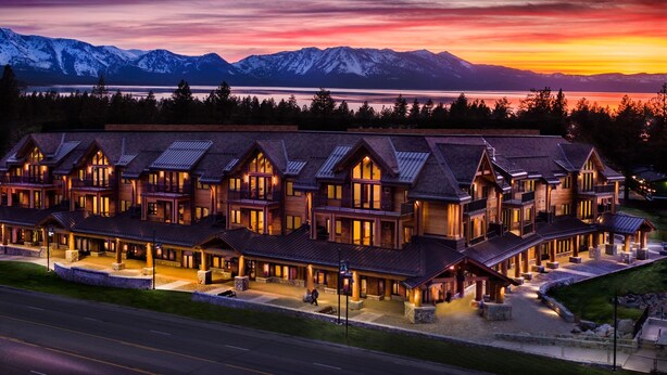 A large, multi-story lodge with a symmetrical roofline and multiple balconies, situated in a mountainous area with a view of snow-capped peaks and a lake.