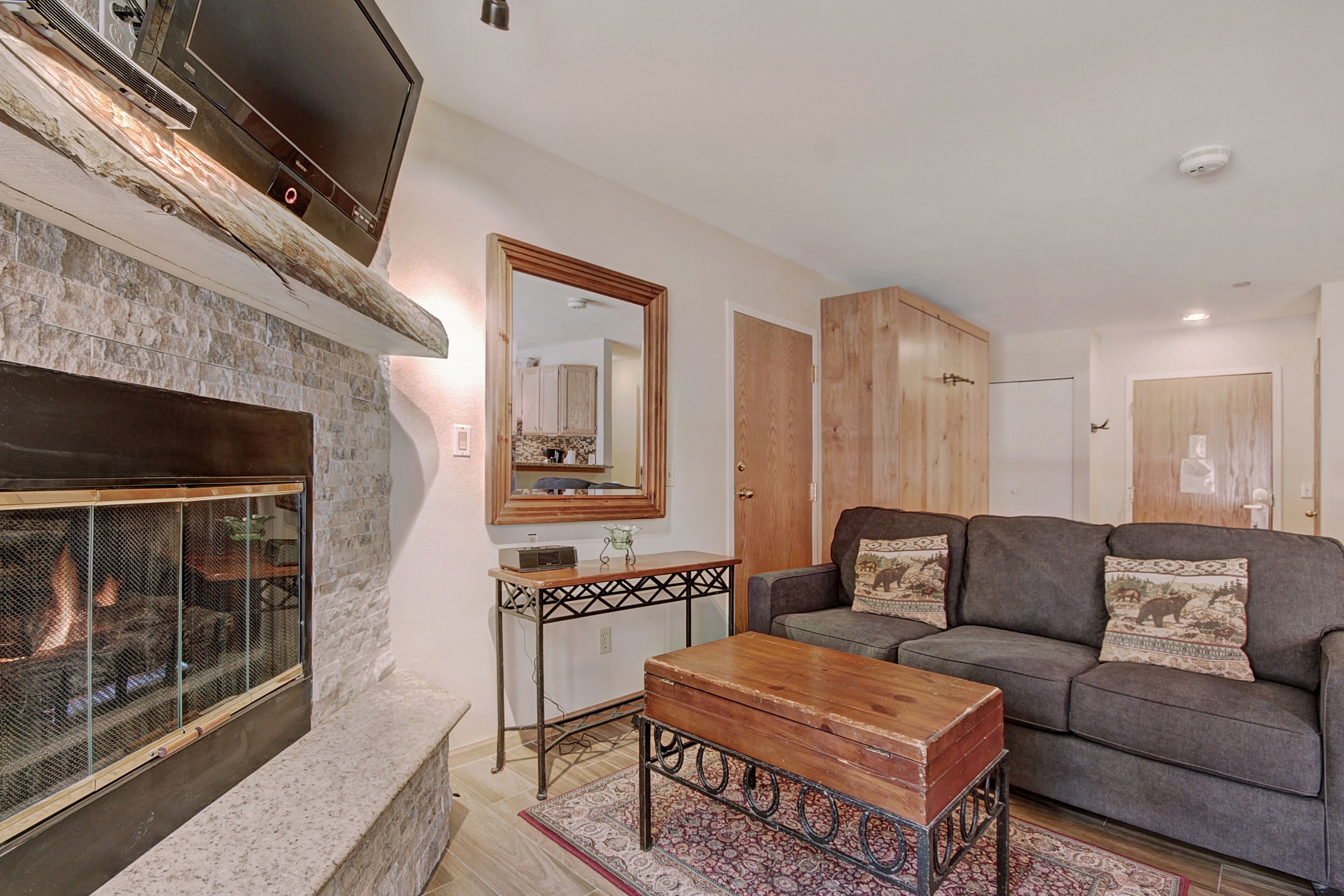 A living room with a stone fireplace, a wooden coffee table, a gray sofa with patterned cushions, and a wooden cabinet.