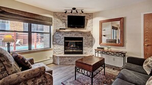 A living room with a stone fireplace, a flat-screen TV mounted above it, a large window with a view of a pool, and a cozy seating area with patterned furniture. - Spacious  In The Heart Of Breckenridge Studio Condo by RedAwning (Breckenridge)