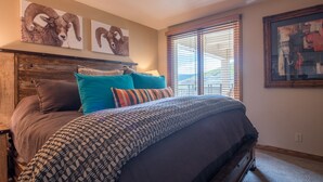 A bedroom with a wooden headboard, a large bed with patterned bedding, and two wall-mounted paintings of rams. - Lovely Modern Plaza Condo - No Cleaning Fee! by RedAwning (Crested Butte)