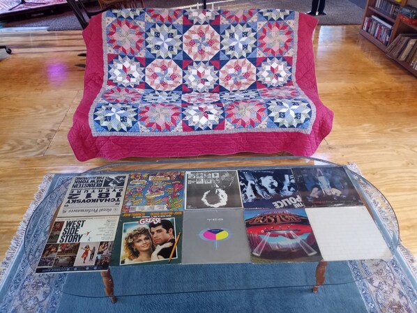 A quilted bedspread with a geometric pattern, a coffee table with magazines and a vinyl record, and a bookshelf in the background.