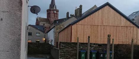 A narrow alleyway with a stone wall on one side and a wooden building with a corrugated metal roof on the other. A stone staircase leads up to a higher level.
