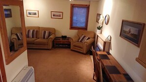 Ein gemütliches Wohnzimmer mit einer Couch, einem Sessel und einem kleinen Holztisch. An der Wand hängt ein Bild in einem Rahmen und eine Uhr steht darauf.