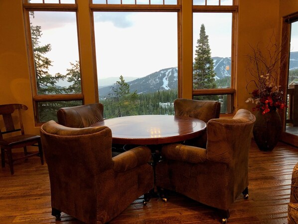 A room with a round wooden table and four leather chairs, large windows with a mountain view, and a vase with red flowers on the right.