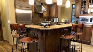 A kitchen with a central island, bar stools, stainless steel appliances, wooden cabinets, and a tiled backsplash. - Moonlight Magic!  Best Views In Big Sky!  True Ski-In/Ski-Out!  Heated Ski Room! (Big Sky)