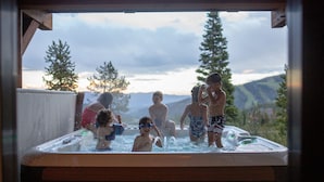 A hot tub with several people around it, surrounded by a wooden deck and overlooking a mountainous landscape. - Moonlight Magic!  Best Views In Big Sky!  True Ski-In/Ski-Out!  Heated Ski Room! (Big Sky)