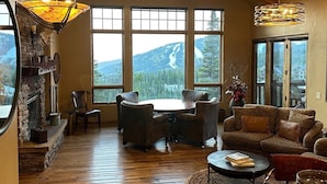 A spacious living room with a stone fireplace, wooden beams, large windows offering mountain views, and a cozy seating area. - Moonlight Magic!  Best Views In Big Sky!  True Ski-In/Ski-Out!  Heated Ski Room! (Big Sky)