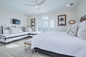 A modern bedroom with a large bed, a sofa bed, a ceiling fan, a TV, and a view of the ocean through a large window.