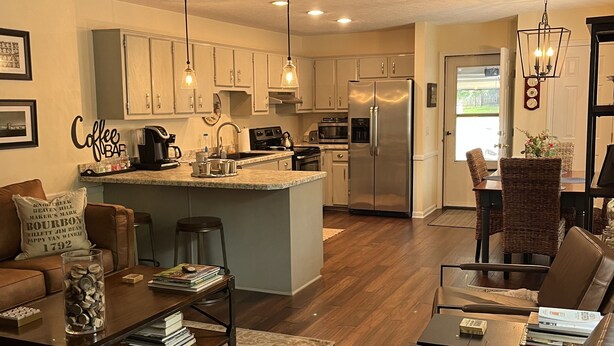 A modern kitchen with a central island, white cabinets, and stainless steel appliances. There is a dining area with wicker chairs and a coffee bar with a sign. - Luxurious 2BR /2Bath with Outdoor Courtyard and Garage in Downtown Bardstown (Bardstown)