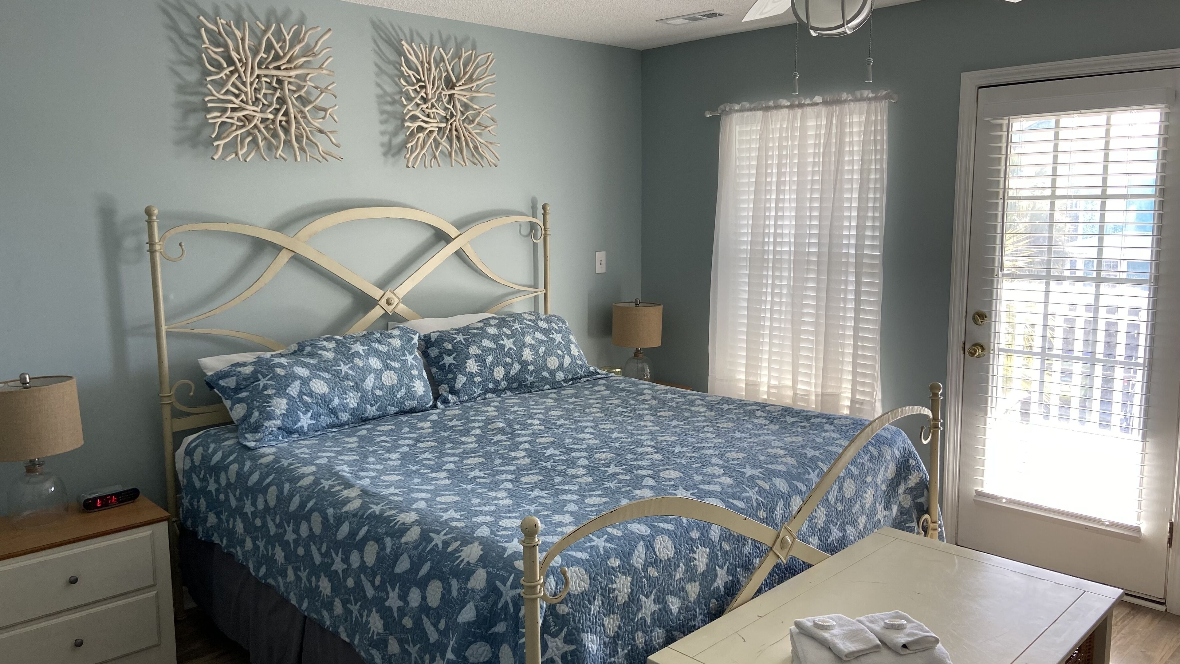 A bedroom with a bed, bedside tables, a nightstand, a lamp, decorative wall art, and a window with blinds.