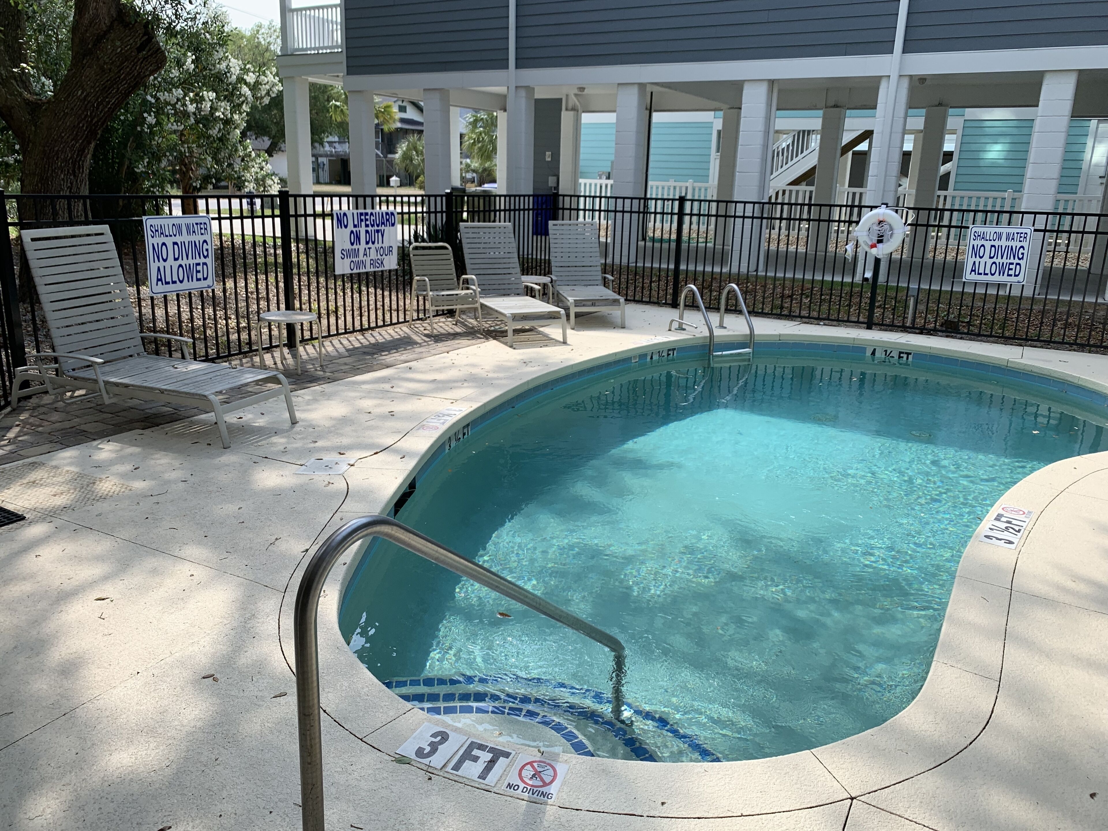 A swimming pool with a 3 ft depth marker, surrounded by lounge chairs and a fence with warning signs.