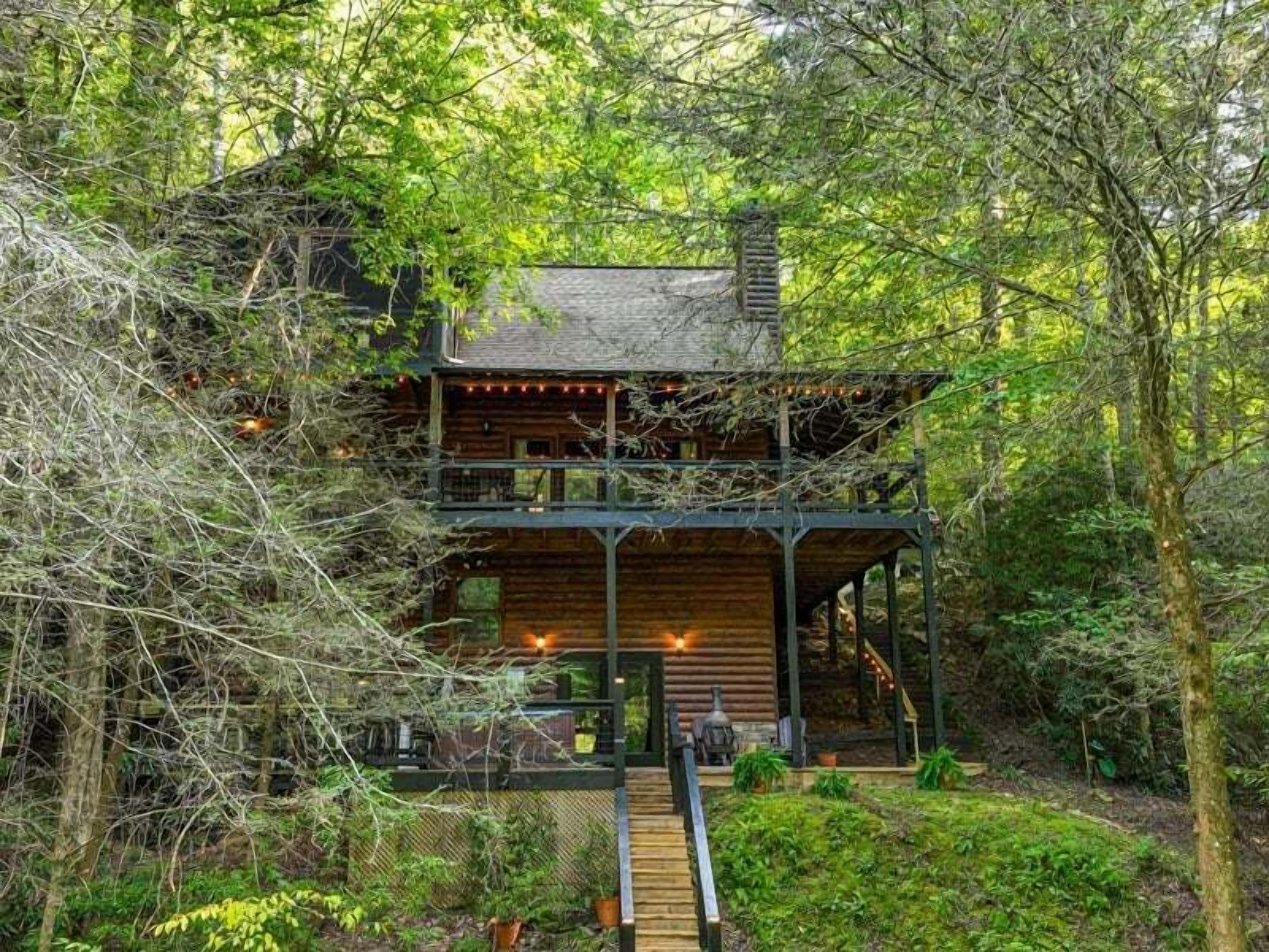 Une cabane en rondins à deux étages, dotée d’un balcon et d’un espace extérieur aménagé pour s’asseoir, entourée d’une forêt dense.