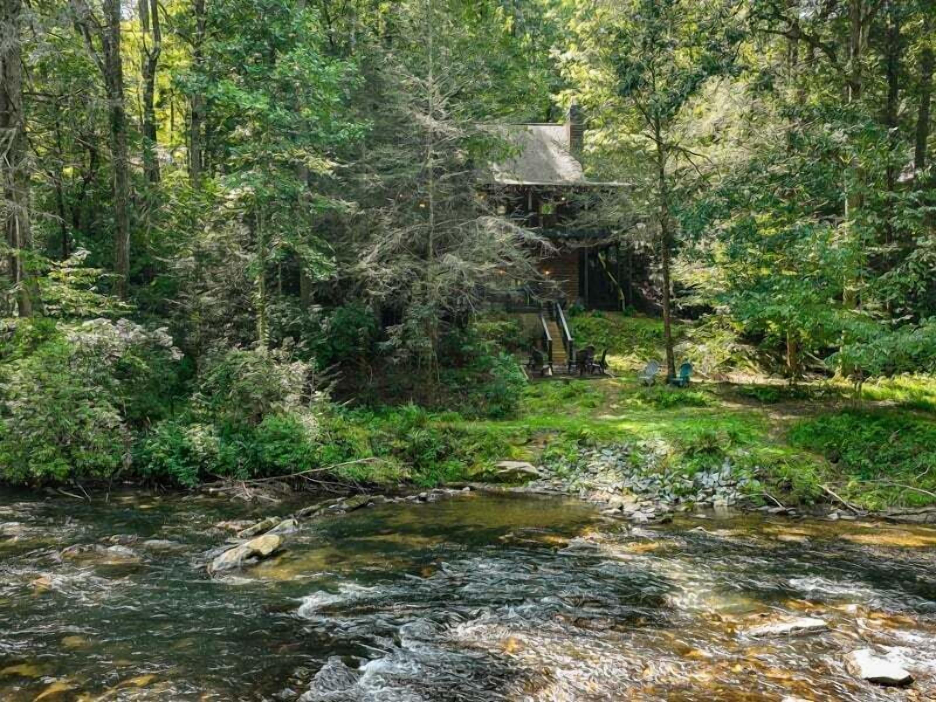 Une cabane entourée d’une forêt dense, avec un petit ruisseau à proximité et une terrasse en bois dotée de sièges d’extérieur.